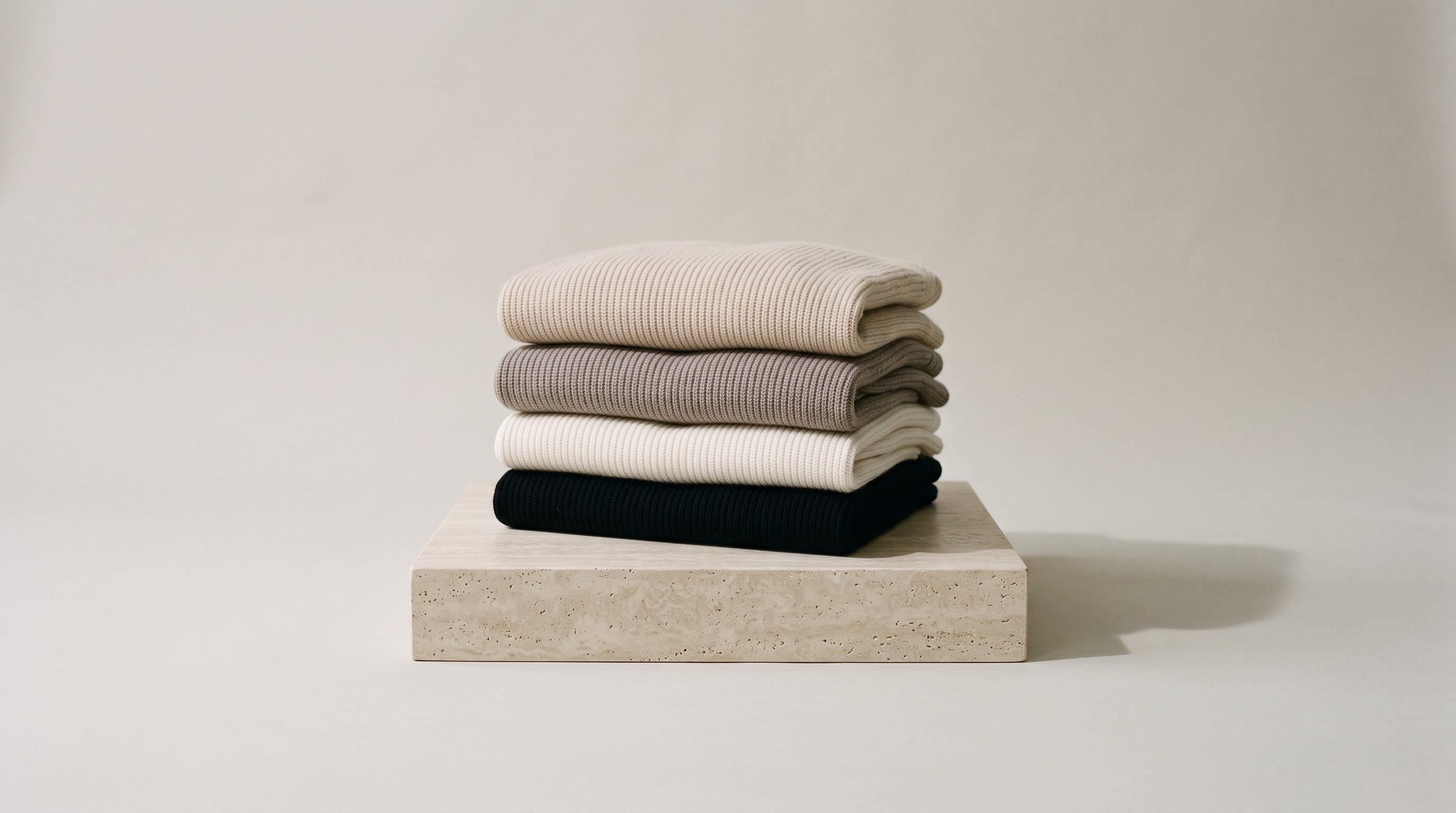 Stacked pure-cashmere knitwear in cream, taupe and oat on a stone plinth, studio backdrop
