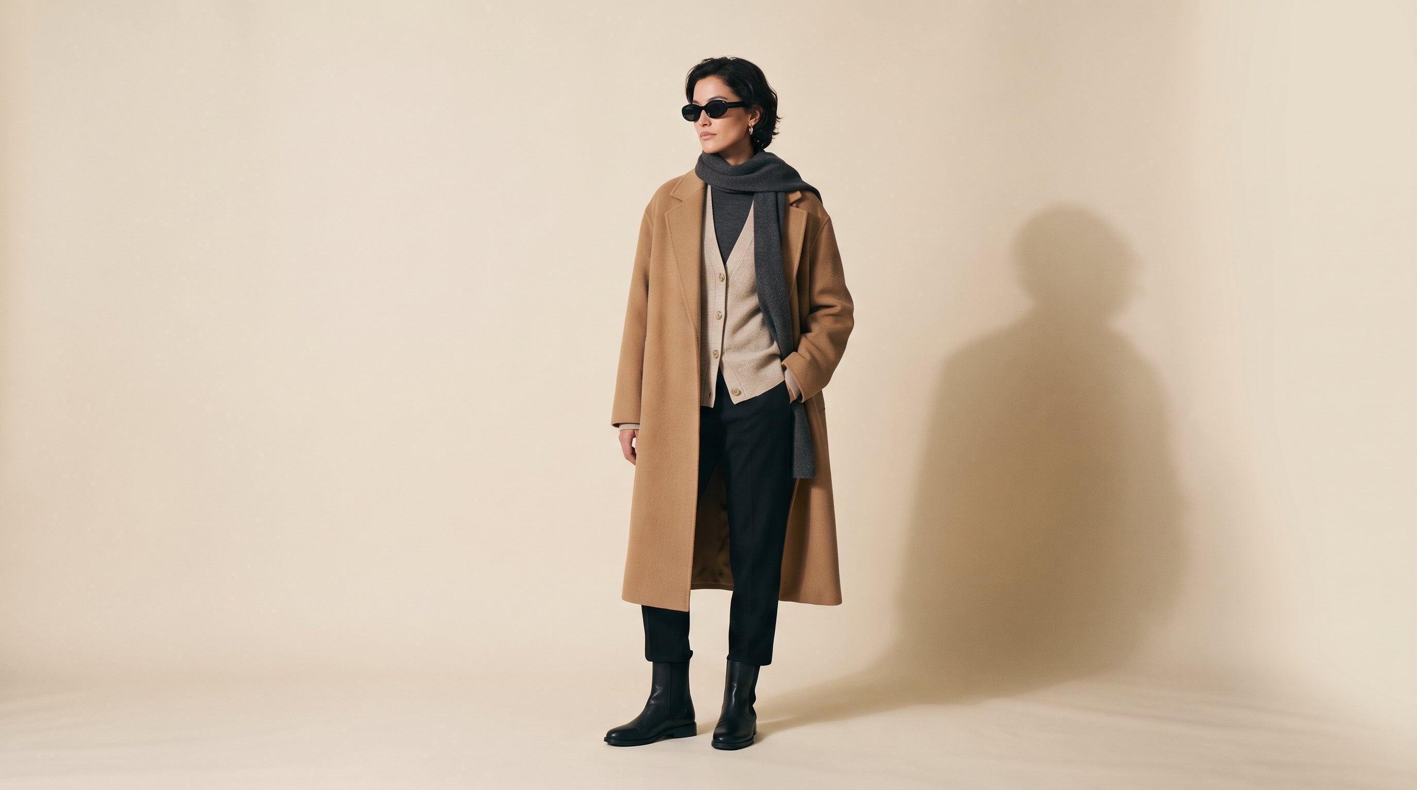 Model in a camel wool overcoat and oat cardigan layered knitwear, studio backdrop